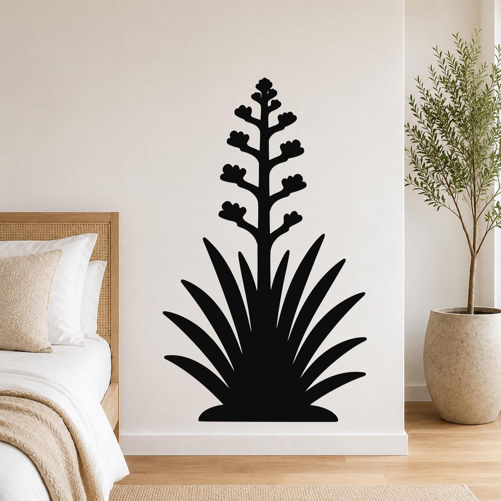 Black metal wall art silhouette of a blooming agave plant with spiky leaves and flower stalk, mounted on a bedroom wall above a bed.