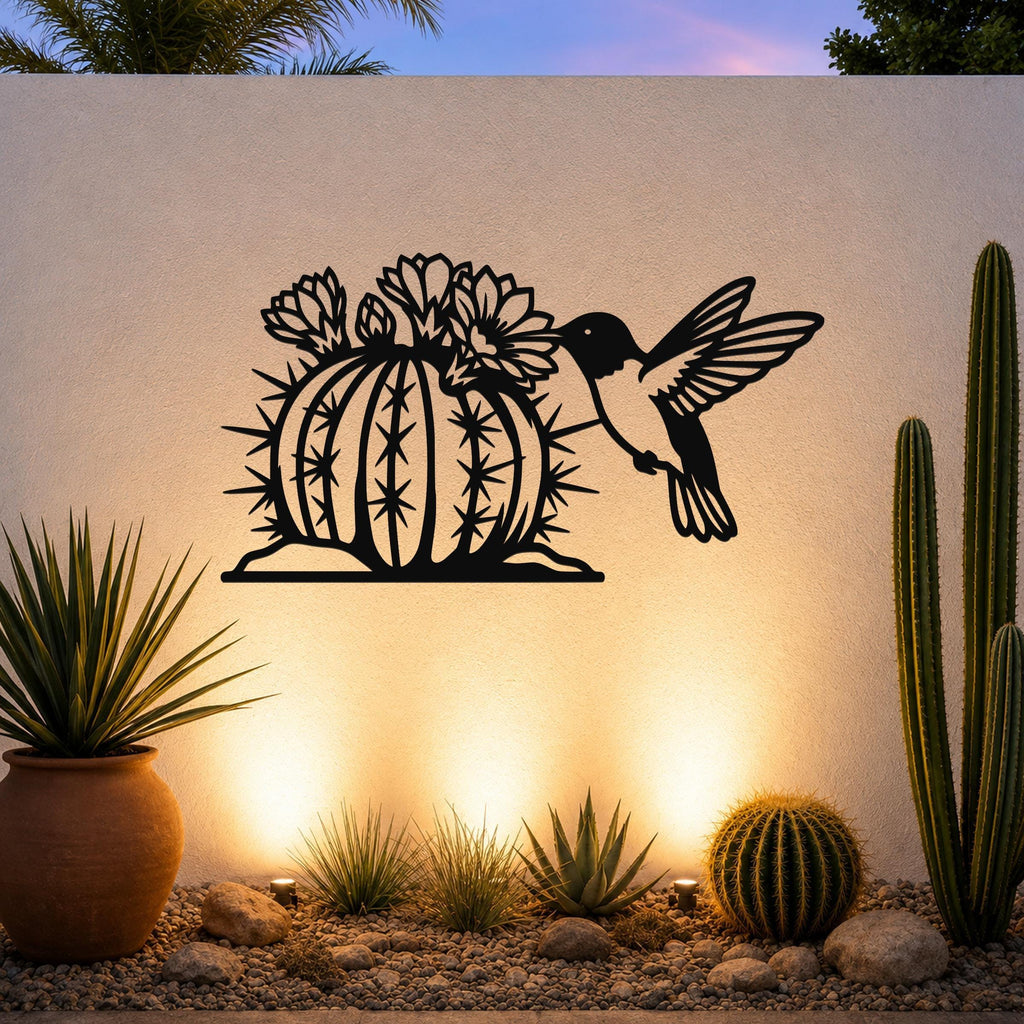 Black metal wall art featuring a hummingbird and flowering barrel cactus cutout, mounted on a white outdoor wall above a desert garden with potted plants and ground lights.