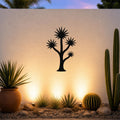 Black metal wall art in a tall yucca silhouette with starburst leaves, mounted on an outdoor wall above a desert garden with cacti and warm uplighting.