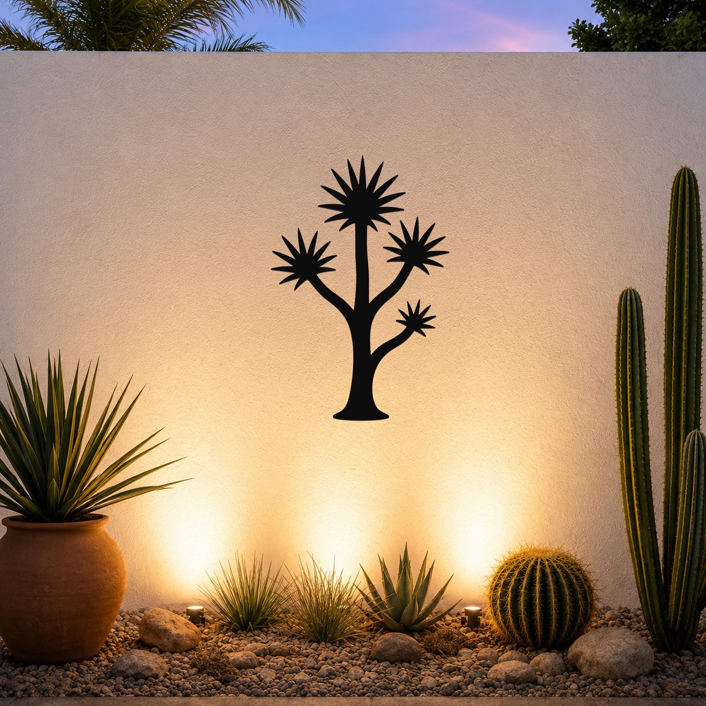 Black metal wall art in a tall yucca silhouette with starburst leaves, mounted on an outdoor wall above a desert garden with cacti and warm uplighting.