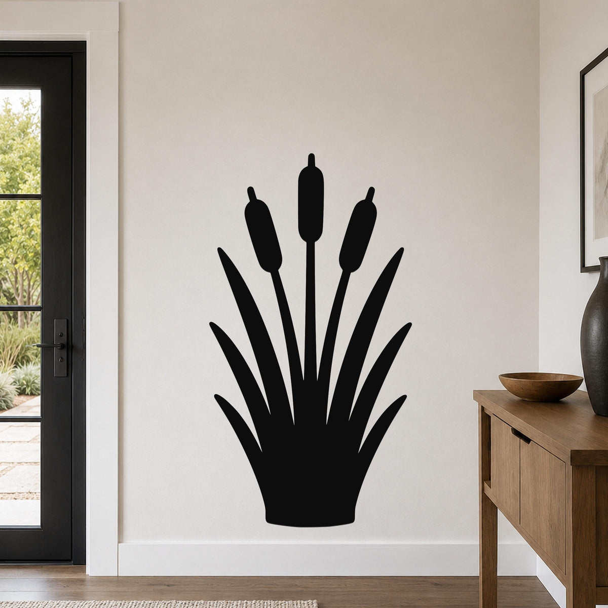 Black metal wall art silhouette of cattails with long leaves and three seed heads, mounted on a white wall in a living room near a door and console table.