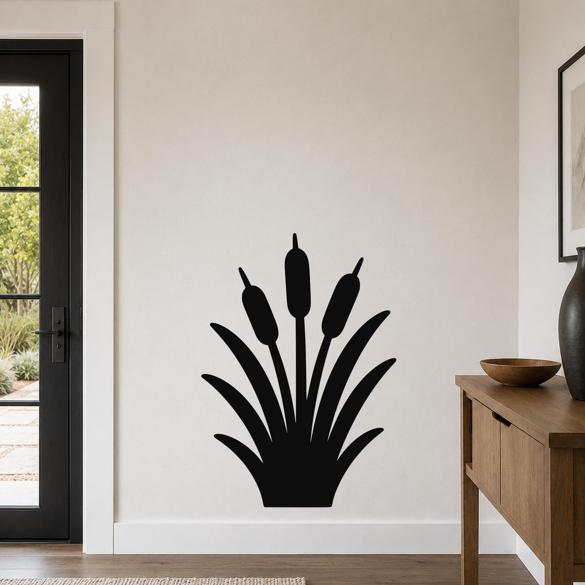 Black metal cattail wall art silhouette with three seed heads and long leaves, mounted on a white wall in a living room near a door and wooden console table.