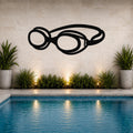 Black metal swim goggles wall art with a bold silhouette design, mounted on a tiled wall above a poolside garden with potted plants and ambient lighting.