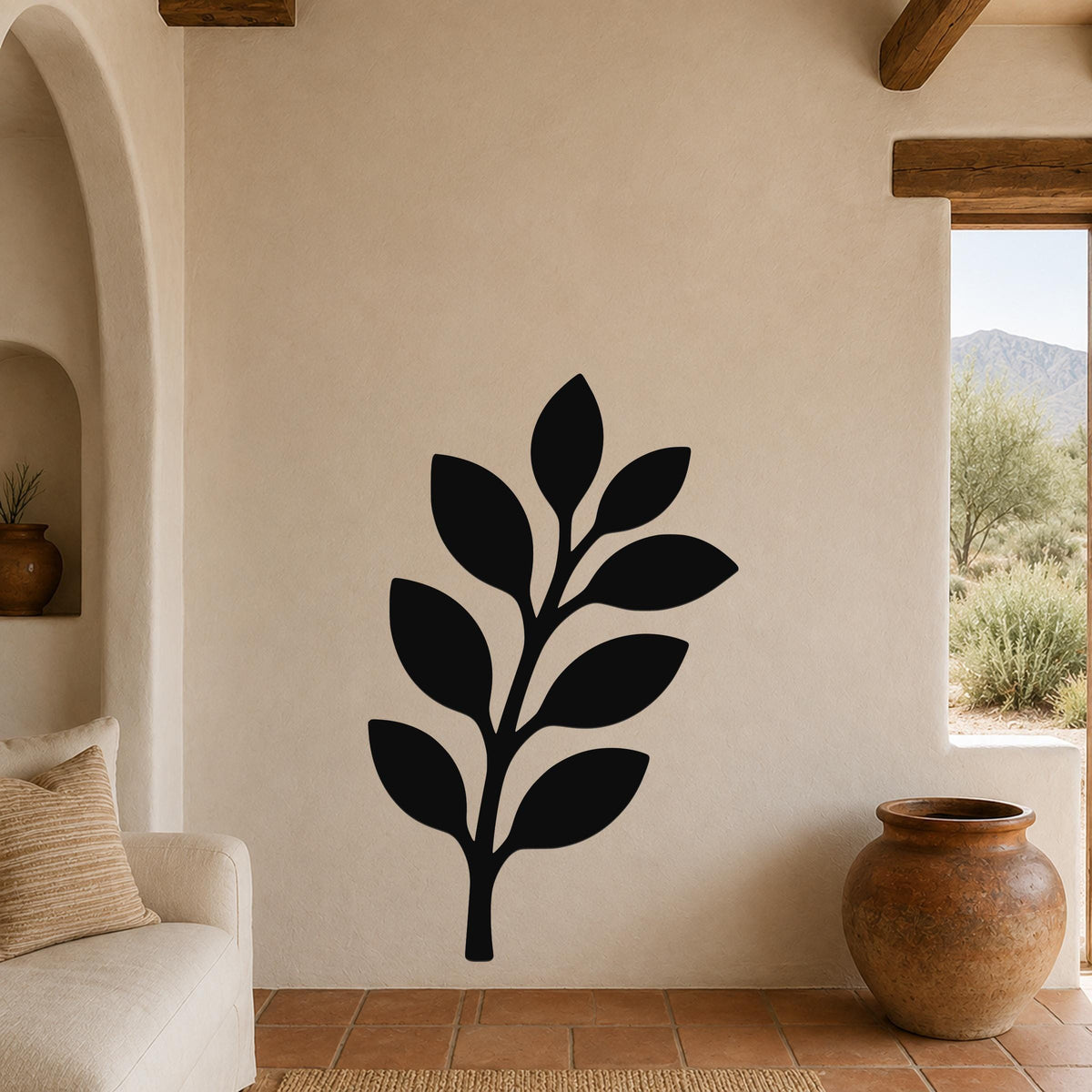 Black metal olive branch wall art with leaf cutouts, mounted on a beige stucco wall in a living room with terracotta flooring and wooden beams.