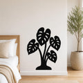 Black metal wall art cutout of a monstera plant with leaf perforations, mounted on a bedroom wall above a bed.