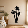 Black metal wall art featuring a silhouette of three palm trees, mounted on a beige bedroom wall above a bed.