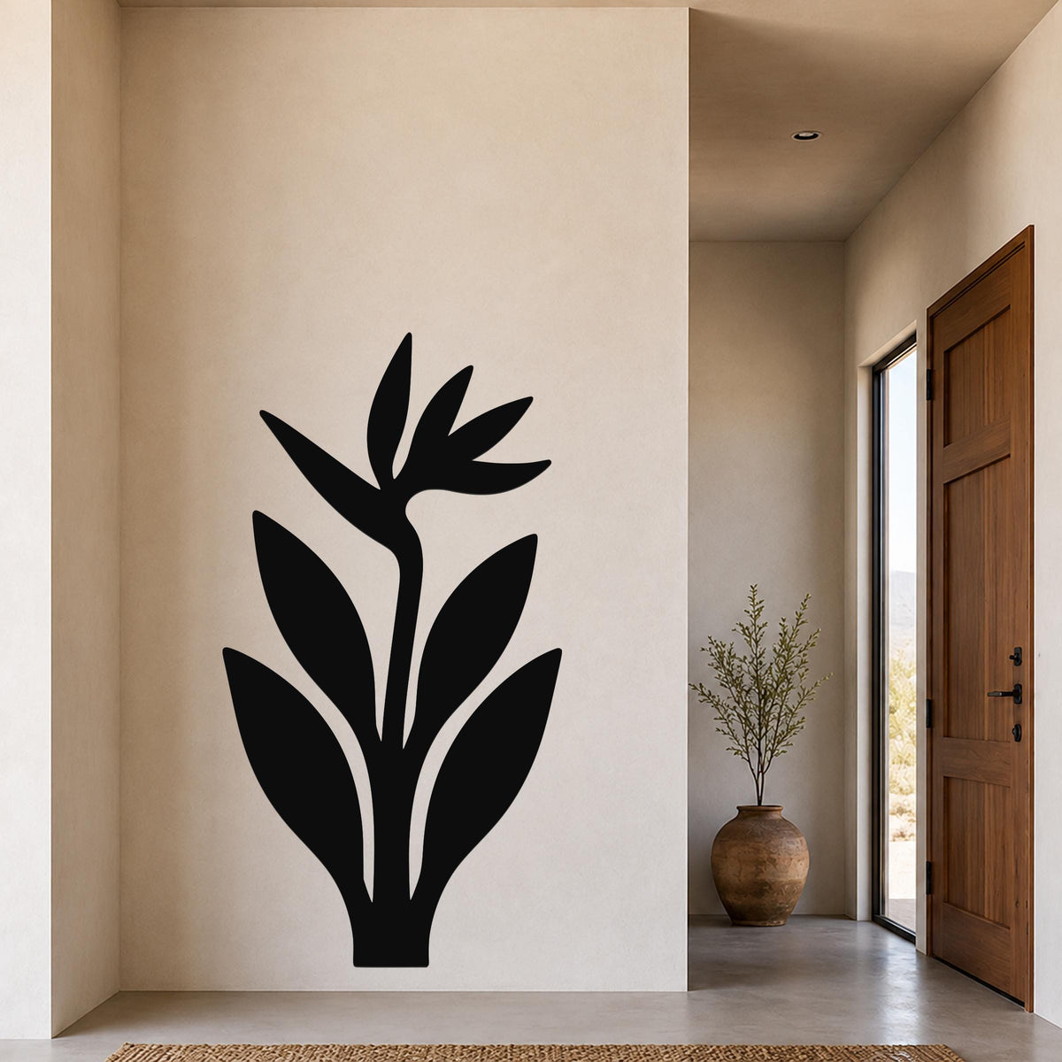 Black metal wall art of a Bird of Paradise plant silhouette, mounted on a beige wall in a modern entryway with a wooden door and a potted plant nearby.