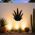 Black metal agave wall art silhouette mounted on a white outdoor wall above a desert garden with cacti and warm uplighting.