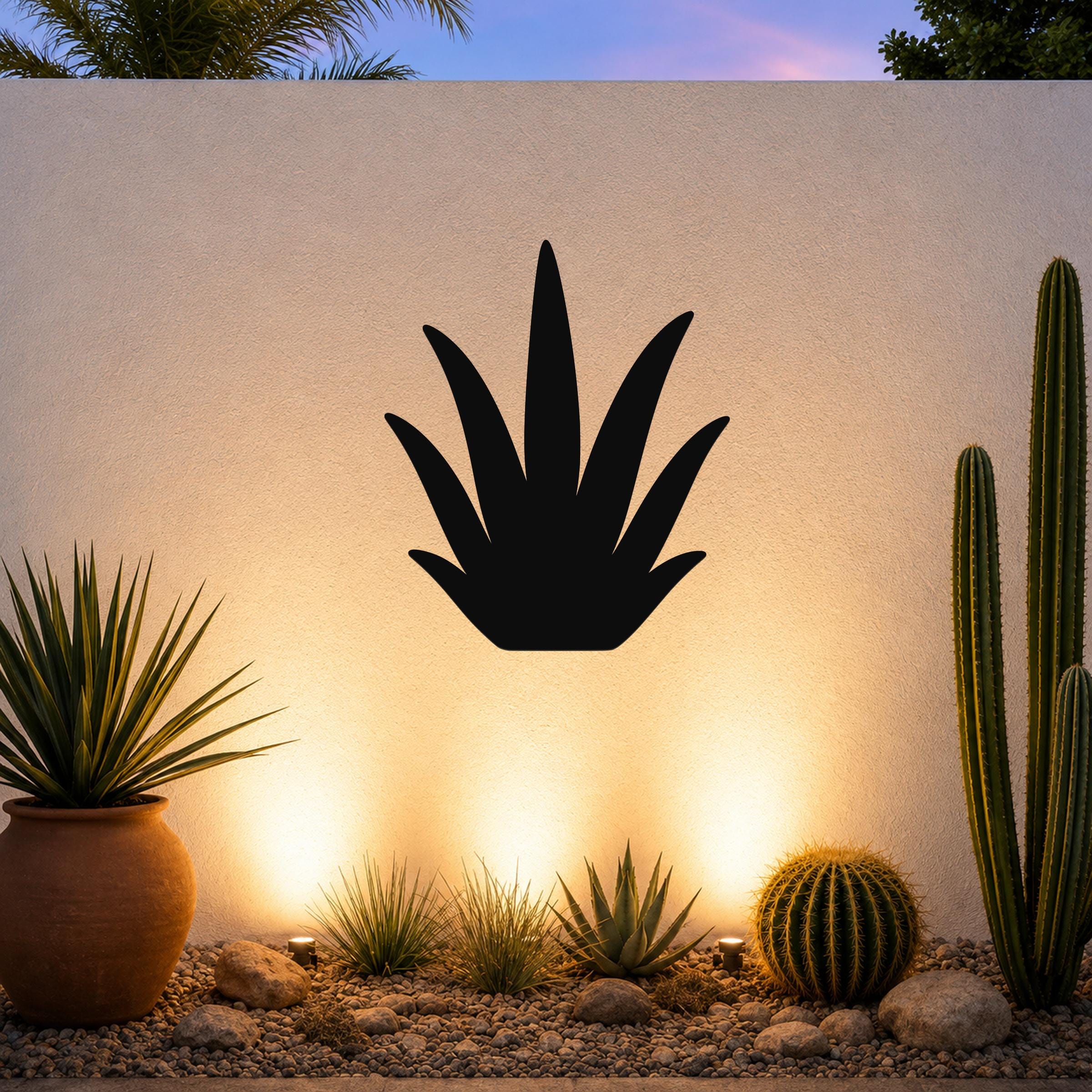 Black metal agave wall art silhouette mounted on a white outdoor wall above a desert garden with cacti and warm uplighting.
