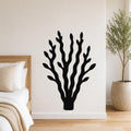 Black metal wall art in a kelp or seaweed silhouette design, mounted on a white bedroom wall above a bed with a woven headboard.