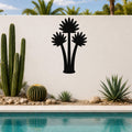 Black metal wall art featuring a fan palm tree silhouette with three fronds, mounted on a white outdoor wall above a pool and desert plants.