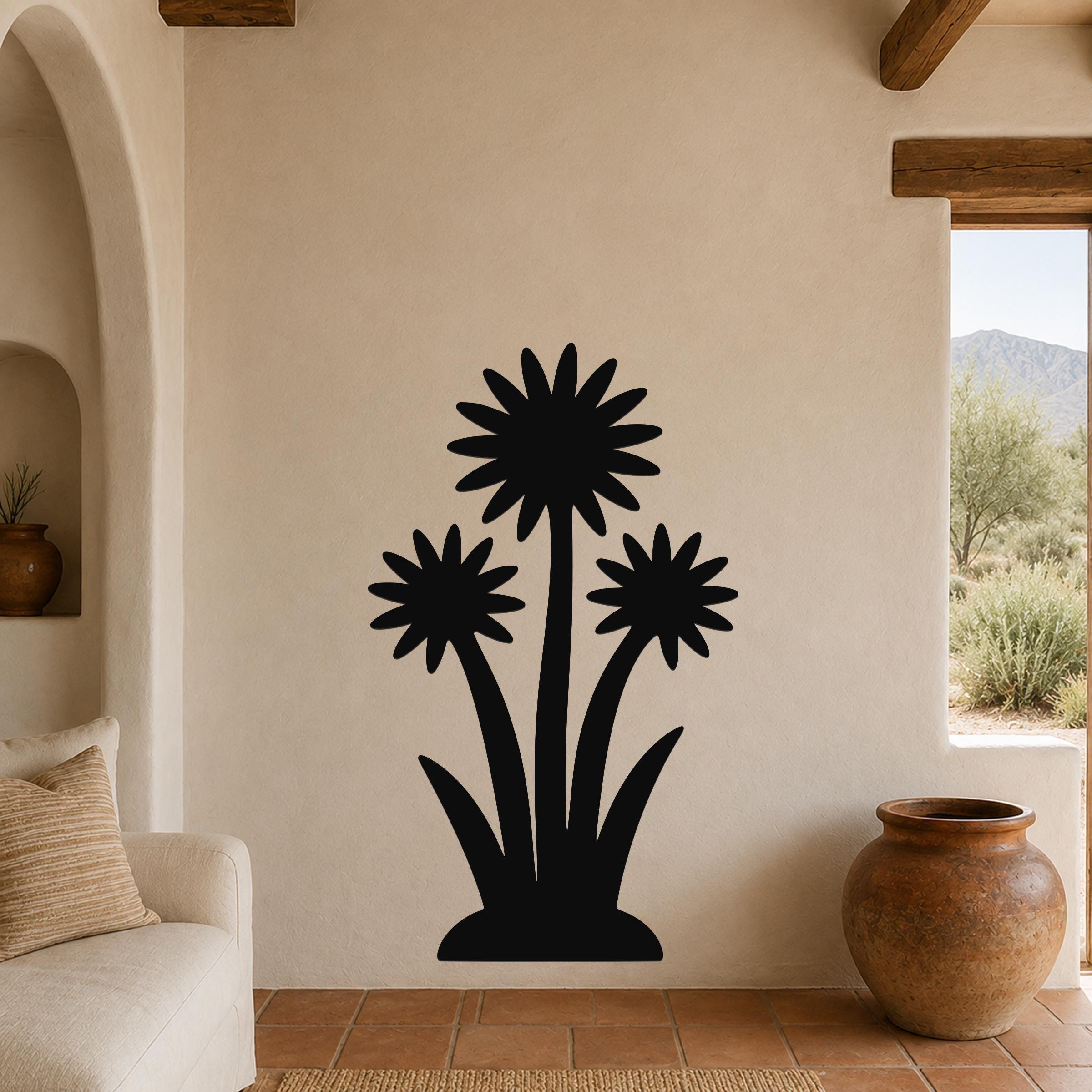 Black metal wall art featuring a silhouette of three wildflowers with pointed petals and long stems, mounted on a beige stucco wall in a living room with terracotta flooring and wooden beams.