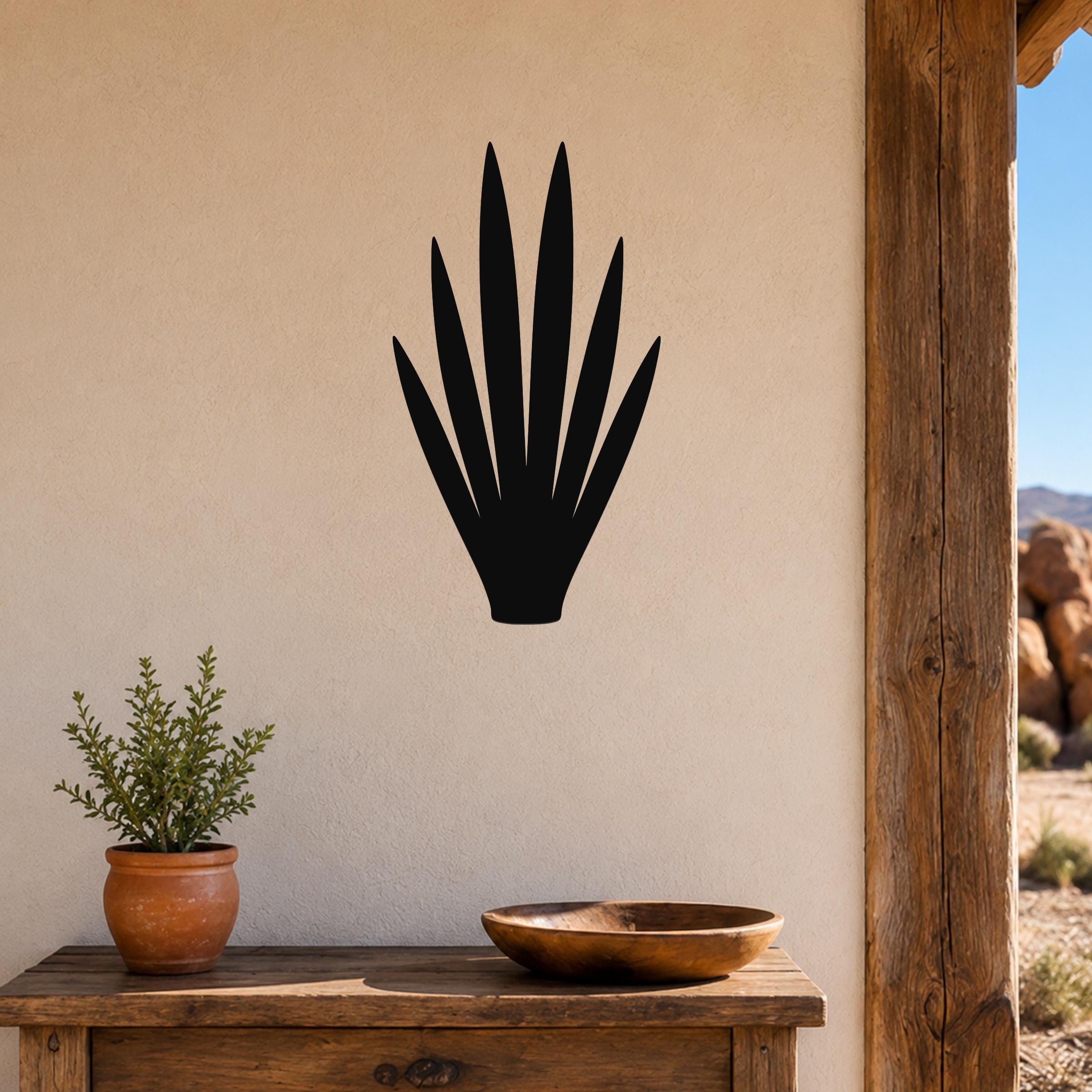 Black metal yucca wall art silhouette mounted on a beige stucco wall above a wooden table in a desert-style patio setting.