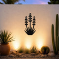 Black metal wall art in a yucca plant silhouette with three tall flower stalks and spiky base, mounted on a textured outdoor wall above a desert garden with cacti and warm uplighting.