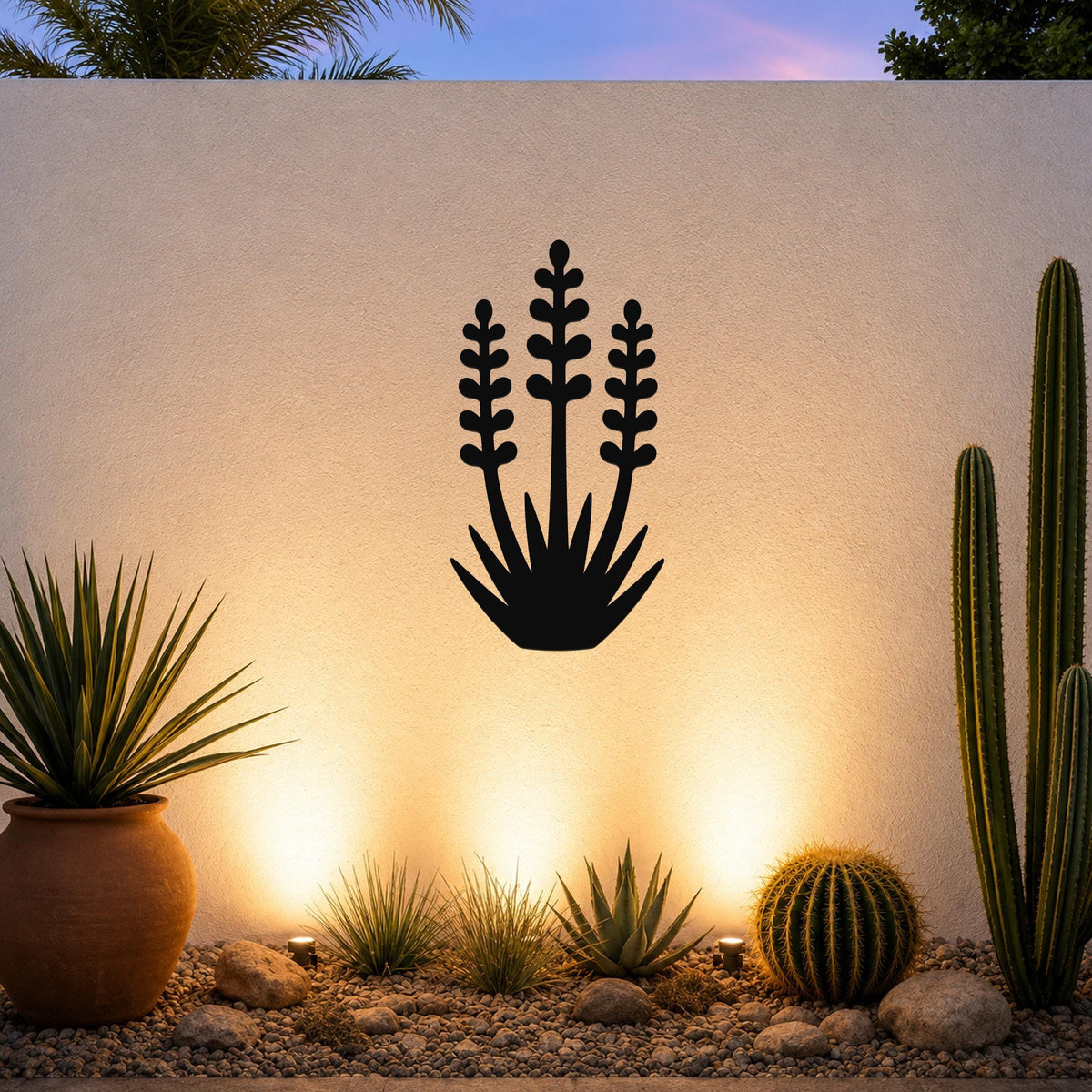 Black metal wall art in a yucca plant silhouette with three tall flower stalks and spiky base, mounted on a textured outdoor wall above a desert garden with cacti and warm uplighting.