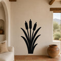 Black metal wall art silhouette of a cattail plant with three seed heads and long leaves, mounted on a beige stucco wall in a living room with terracotta flooring and wooden beams.