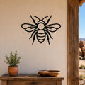 Black metal honey bee wall art with detailed cutout wings and stripes, mounted on a beige stucco wall above a wooden table in a patio setting.