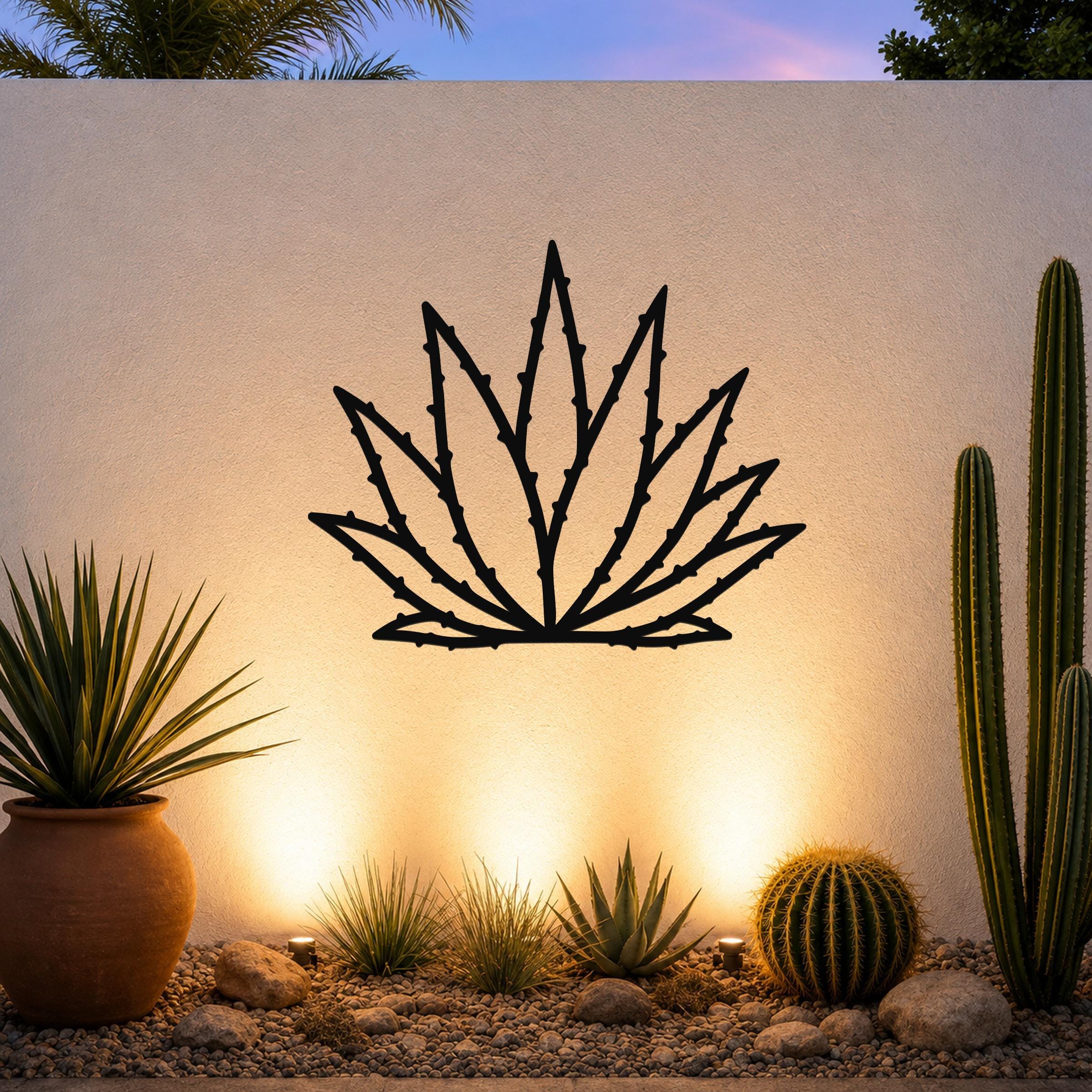 Black metal agave wall art with spiky leaf cutouts, mounted on a textured outdoor wall above a desert garden with cacti and warm uplighting.