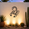 Black metal scorpion wall art with detailed cutout design, mounted on a textured outdoor wall above a desert garden with cacti and warm uplighting.