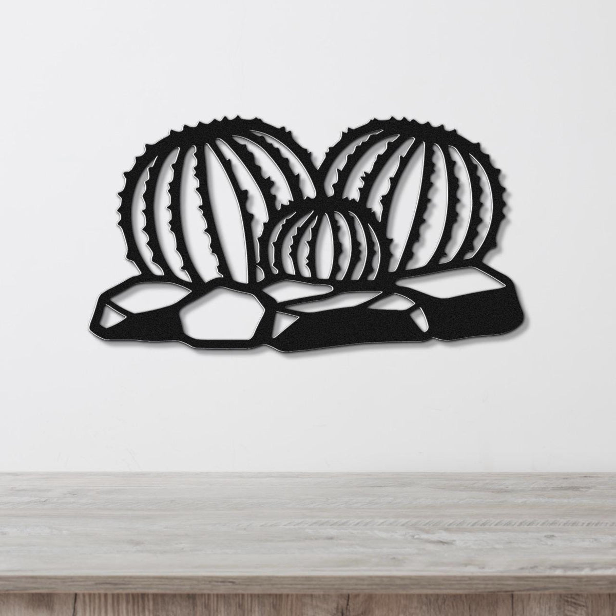 Black metal barrel cactus wall art with ribbed cutout design, mounted on a white wall above a wooden surface.