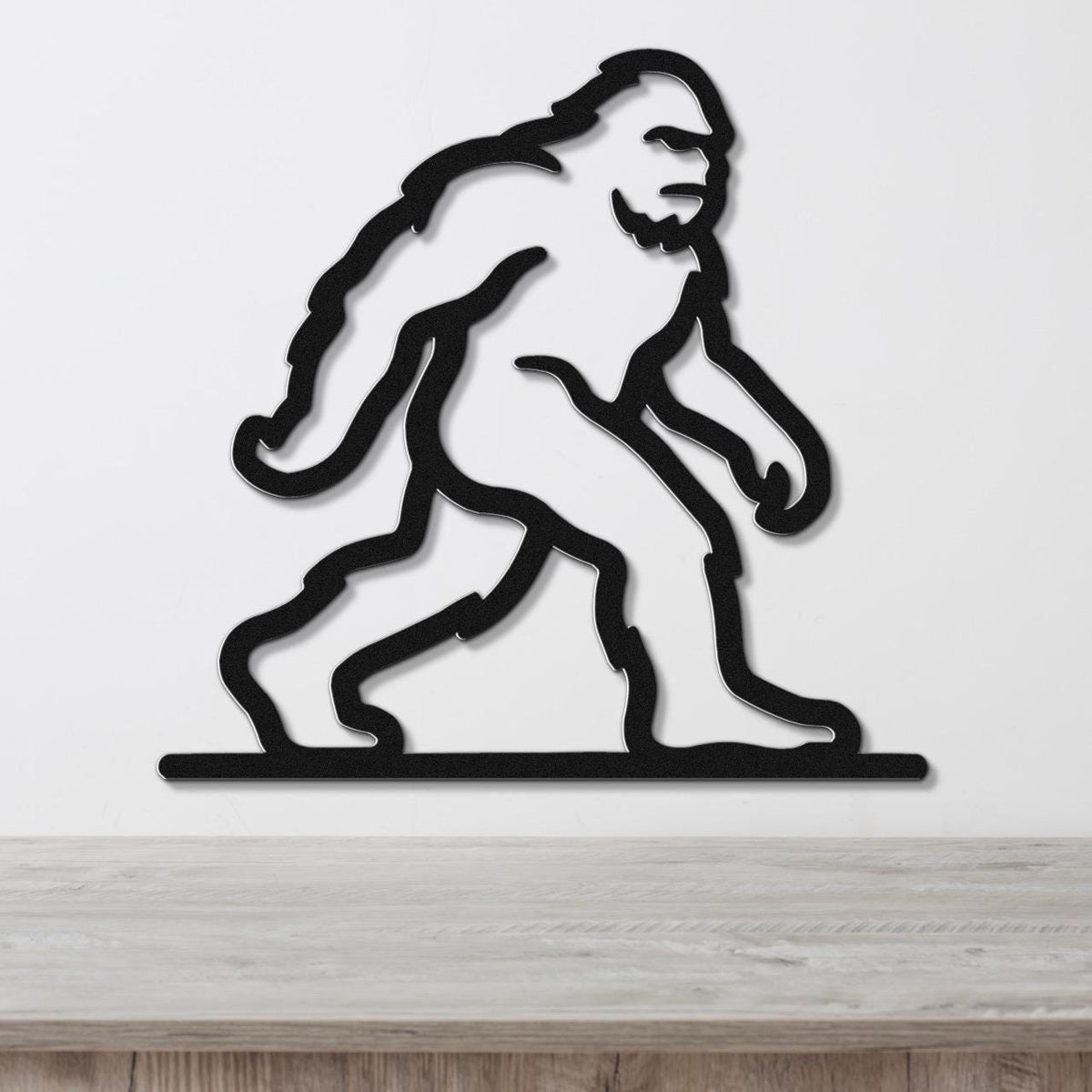 Black metal wall art silhouette of Bigfoot in a walking pose, mounted on a white wall above a wooden surface.