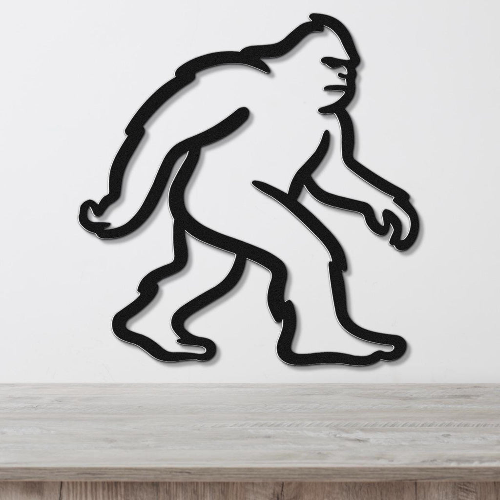 Black metal wall art featuring a minimalist Bigfoot silhouette, mounted on a white wall above a wooden surface.