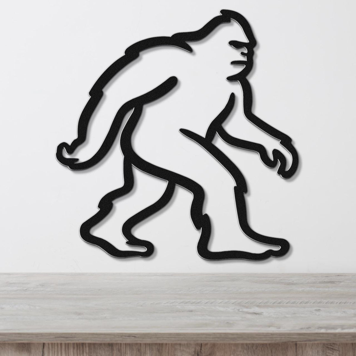Black metal wall art featuring a minimalist Bigfoot silhouette, mounted on a white wall above a wooden surface.