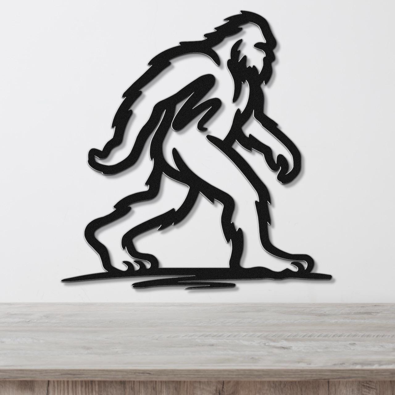 Black metal wall art silhouette of a Bigfoot figure in a walking pose, mounted on a white wall above a wooden surface.