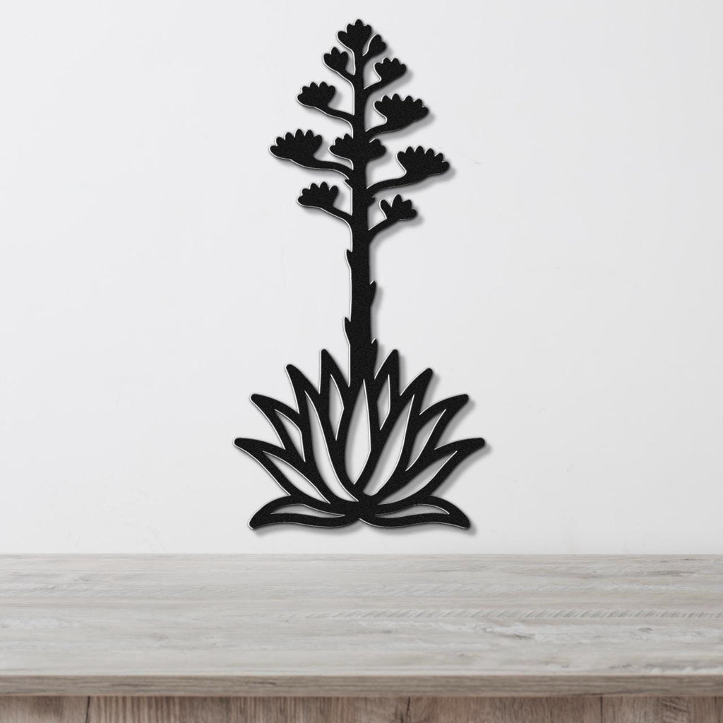 Black metal wall art in the silhouette of a blooming agave plant with a tall flower stalk and spiky rosette base, mounted on a white wall above a light wood surface.
