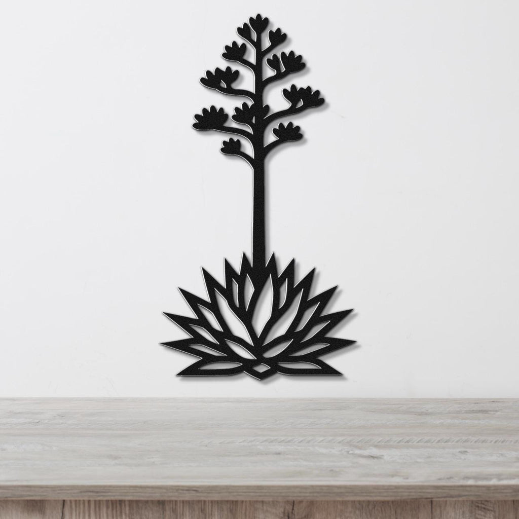 Black metal wall art cutout of a blooming agave plant with a tall stem and flower spikes, mounted on a white wall above a light wood surface.