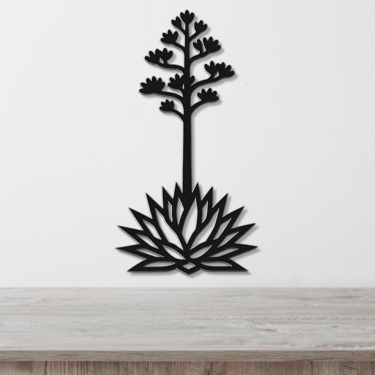 Black metal wall art cutout of a blooming agave plant with a tall stem and flower spikes, mounted on a white wall above a light wood surface.