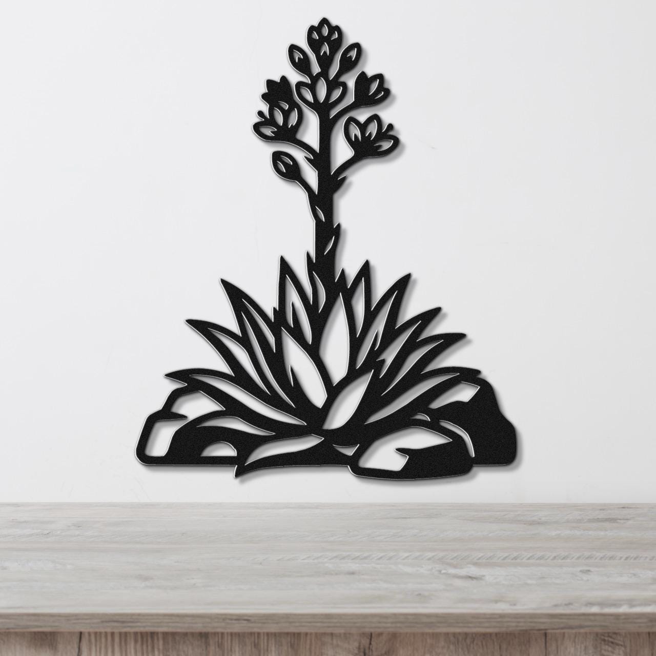 Black metal wall art in a cutout silhouette of a blooming agave plant with spiky leaves and flower stalk, mounted on a white wall above a wooden surface.