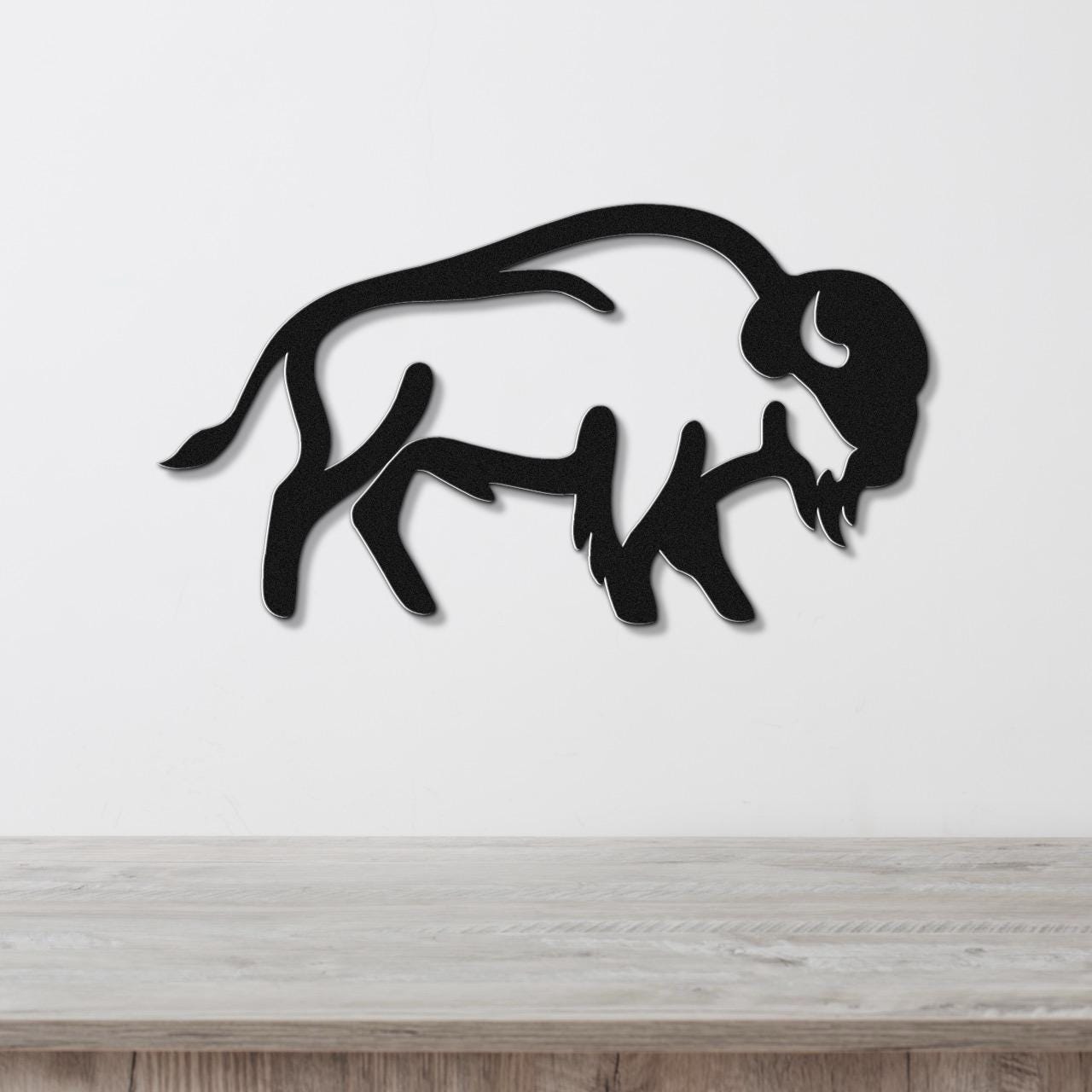 Black metal buffalo silhouette wall art with internal cutouts, mounted on a white wall above a light wood surface.