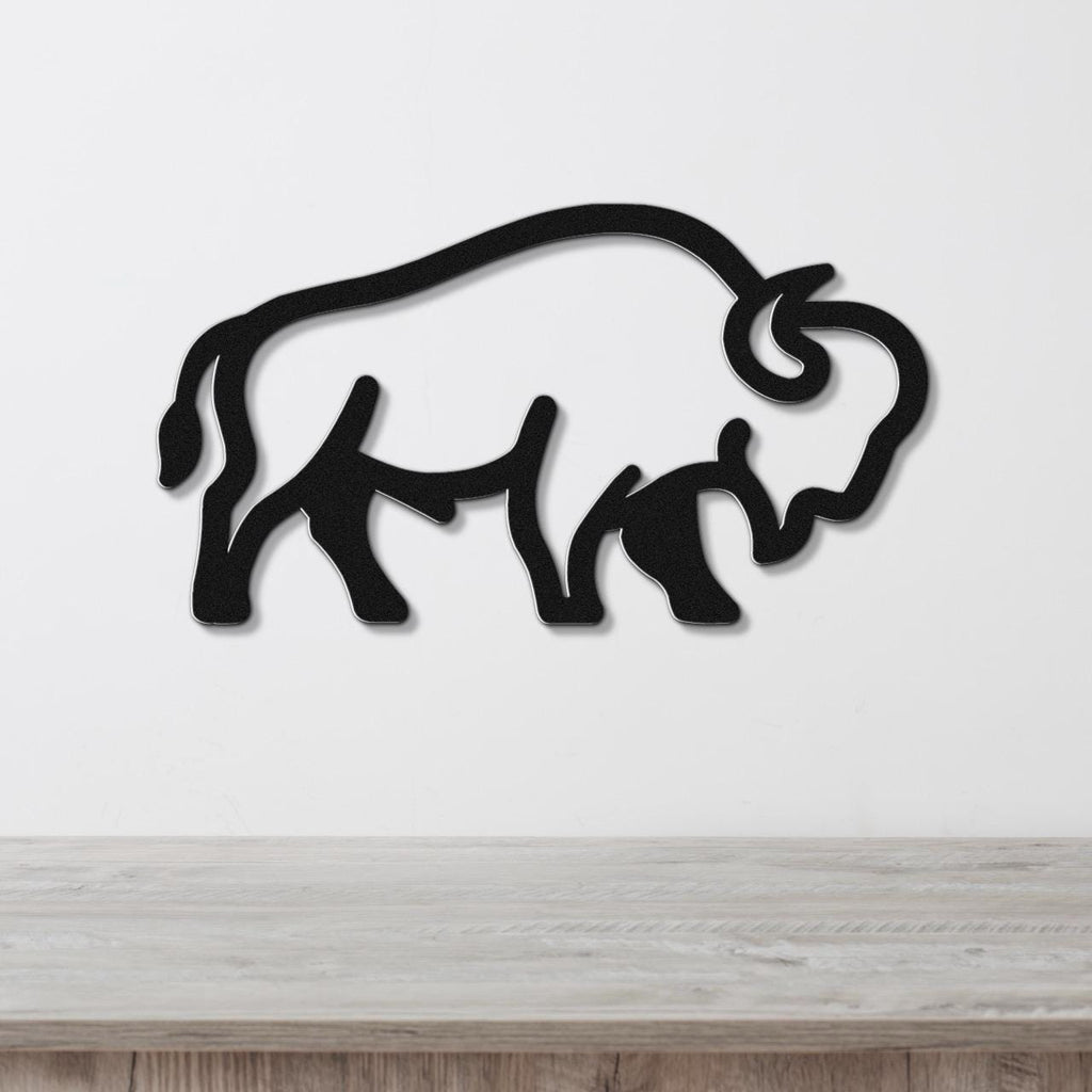 Black metal buffalo silhouette wall art with internal cutout details, mounted on a white wall above a light wood surface.