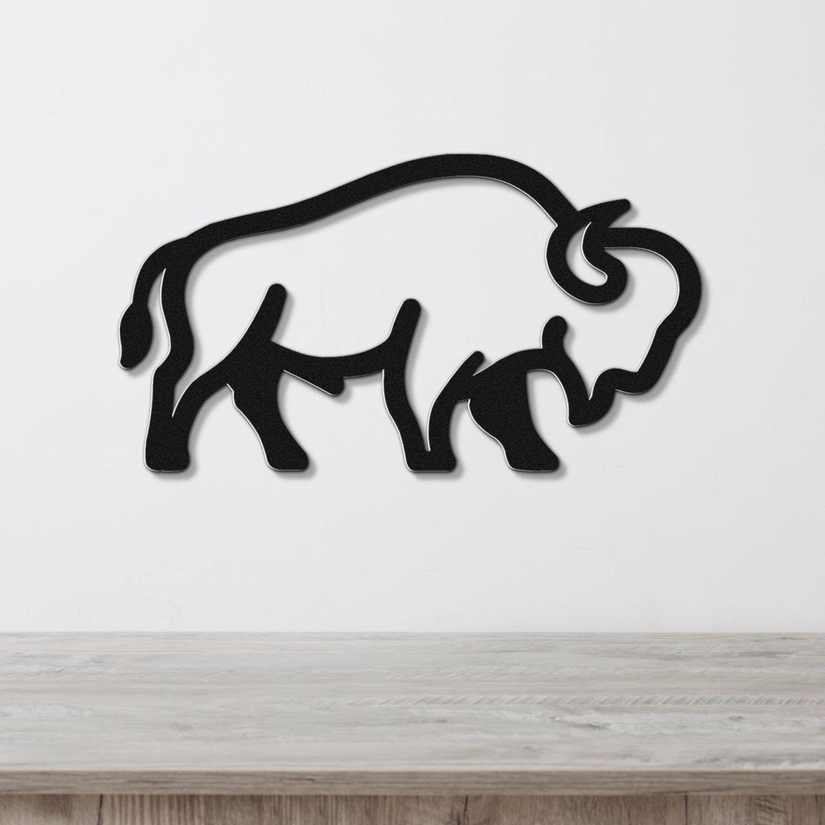 Black metal buffalo silhouette wall art with internal cutout details, mounted on a white wall above a light wood surface.