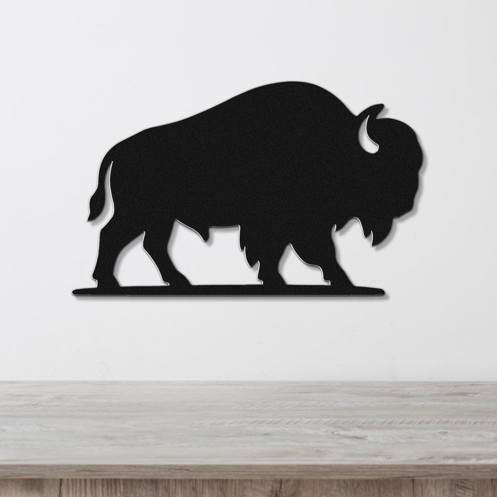 Black metal bison silhouette wall art with cutout horns and legs, mounted on a white wall above a wooden surface.