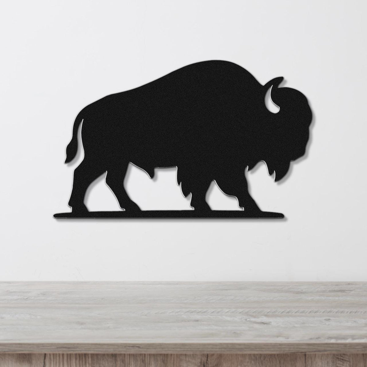 Black metal bison silhouette wall art with cutout horns and legs, mounted on a white wall above a wooden surface.