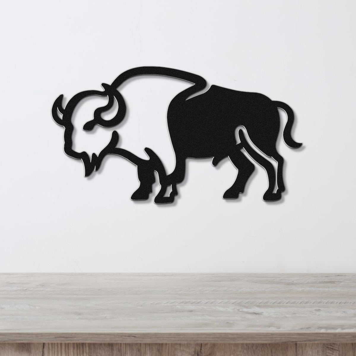 Black metal buffalo wall art silhouette with cutout horns and mane, mounted on a white wall above a light wood surface.