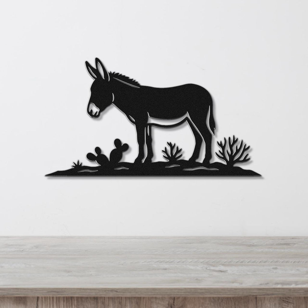 Black metal wall art silhouette of a burro standing in a desert scene with cacti, mounted on a white wall above a wooden surface.