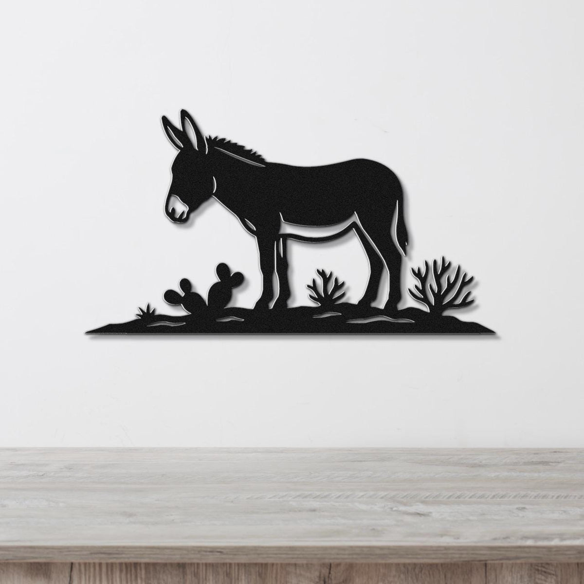 Black metal wall art silhouette of a burro standing in a desert scene with cacti, mounted on a white wall above a wooden surface.