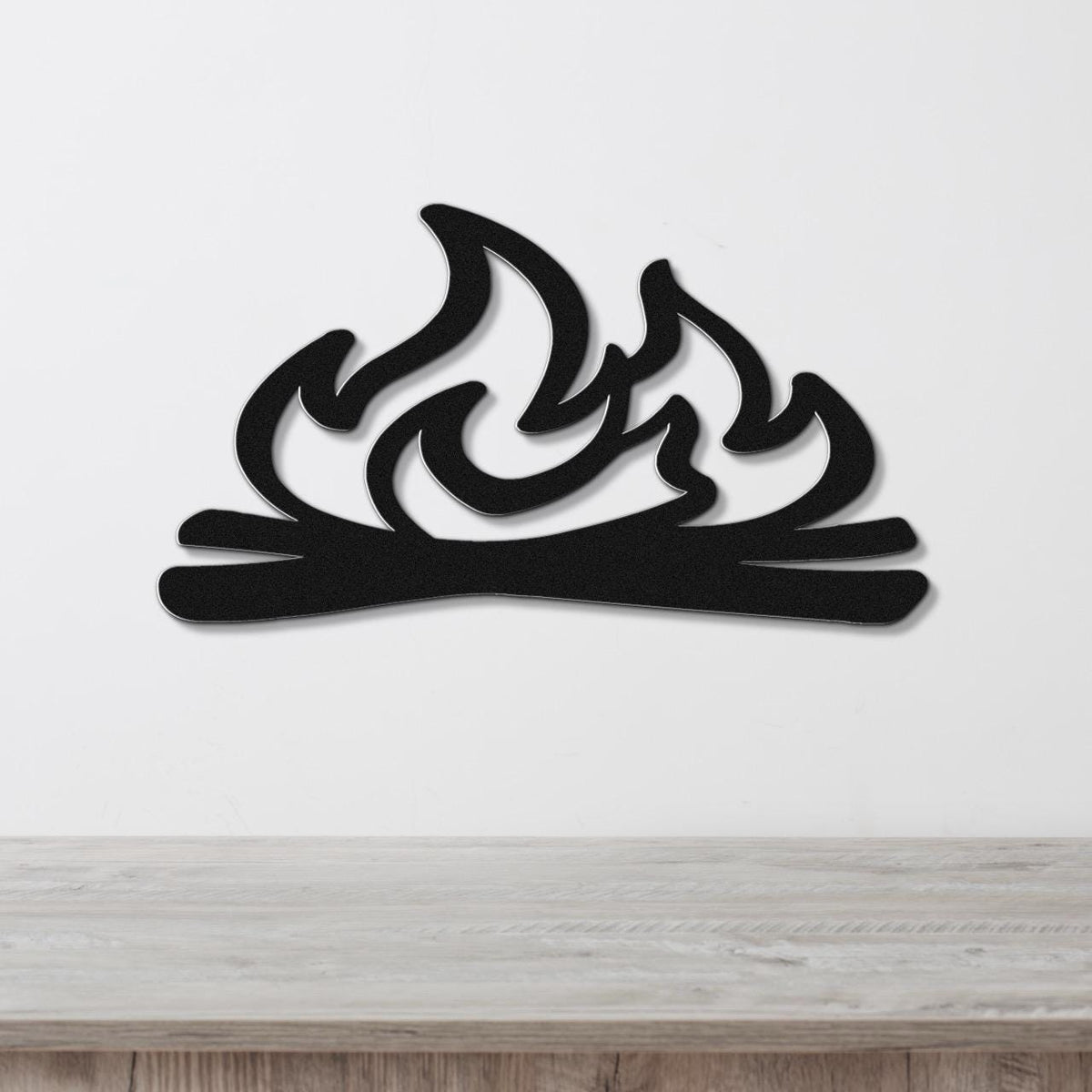 Black metal wall art with a flame and log silhouette, mounted on a white wall above a light wood surface.