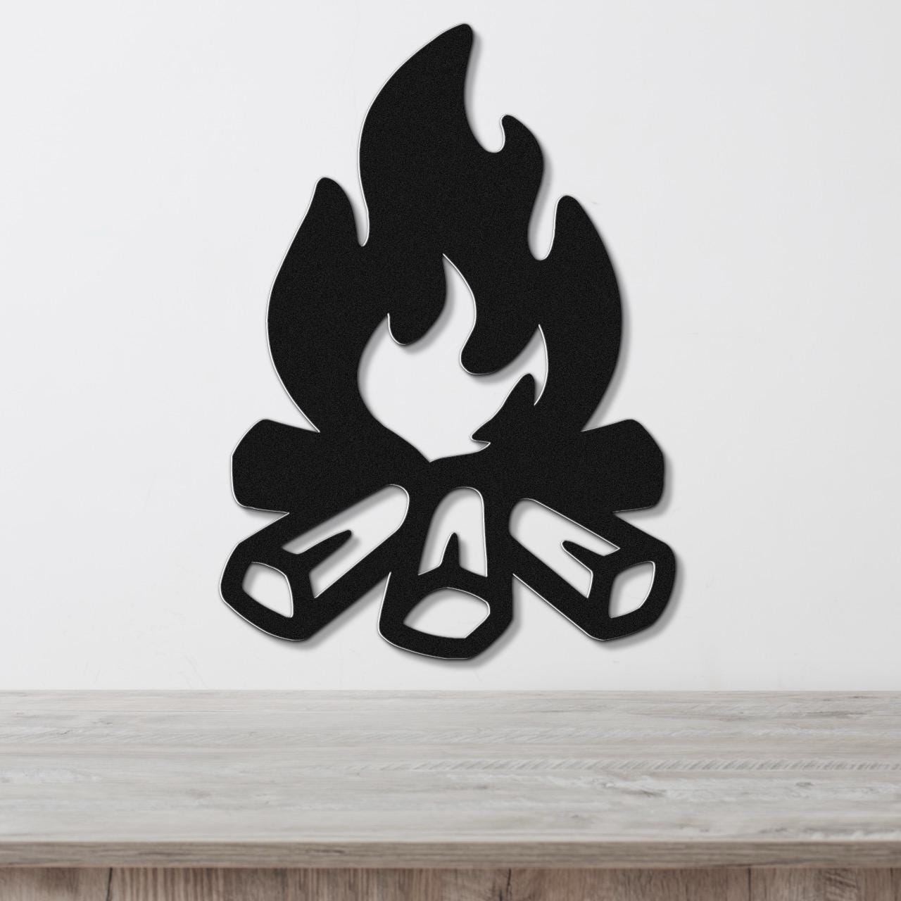 Black metal wall art in a campfire silhouette with flame and log cutouts, mounted on a white wall above a wooden surface.