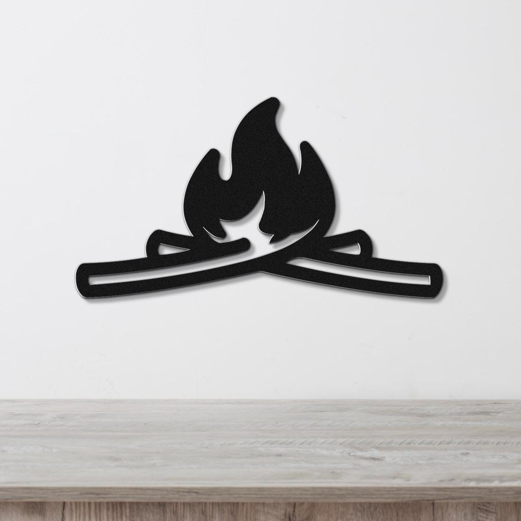 Black metal wall art in a campfire silhouette with crossed logs and flame cutouts, mounted on a white wall above a light wood surface.