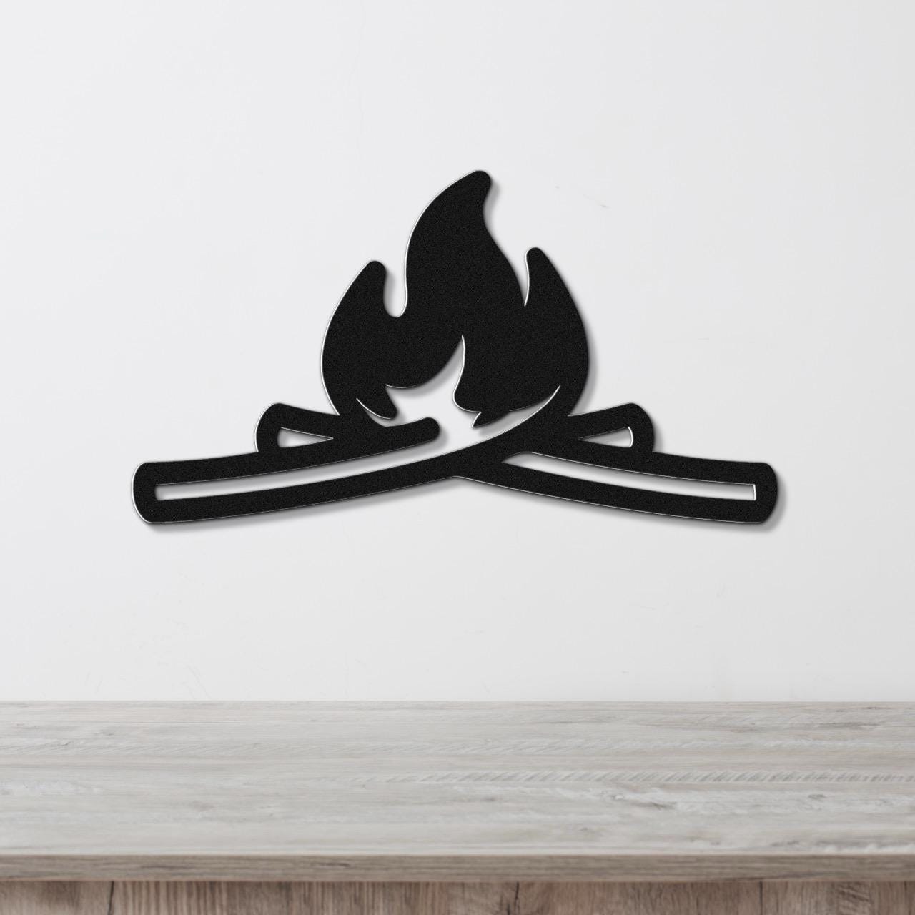 Black metal wall art in a campfire silhouette with crossed logs and flame cutouts, mounted on a white wall above a light wood surface.