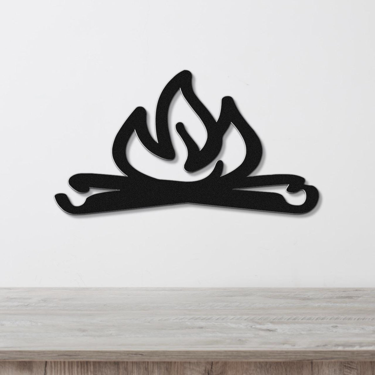 Black metal wall art cutout of a campfire with flames and logs, mounted on a white wall above a wooden surface.