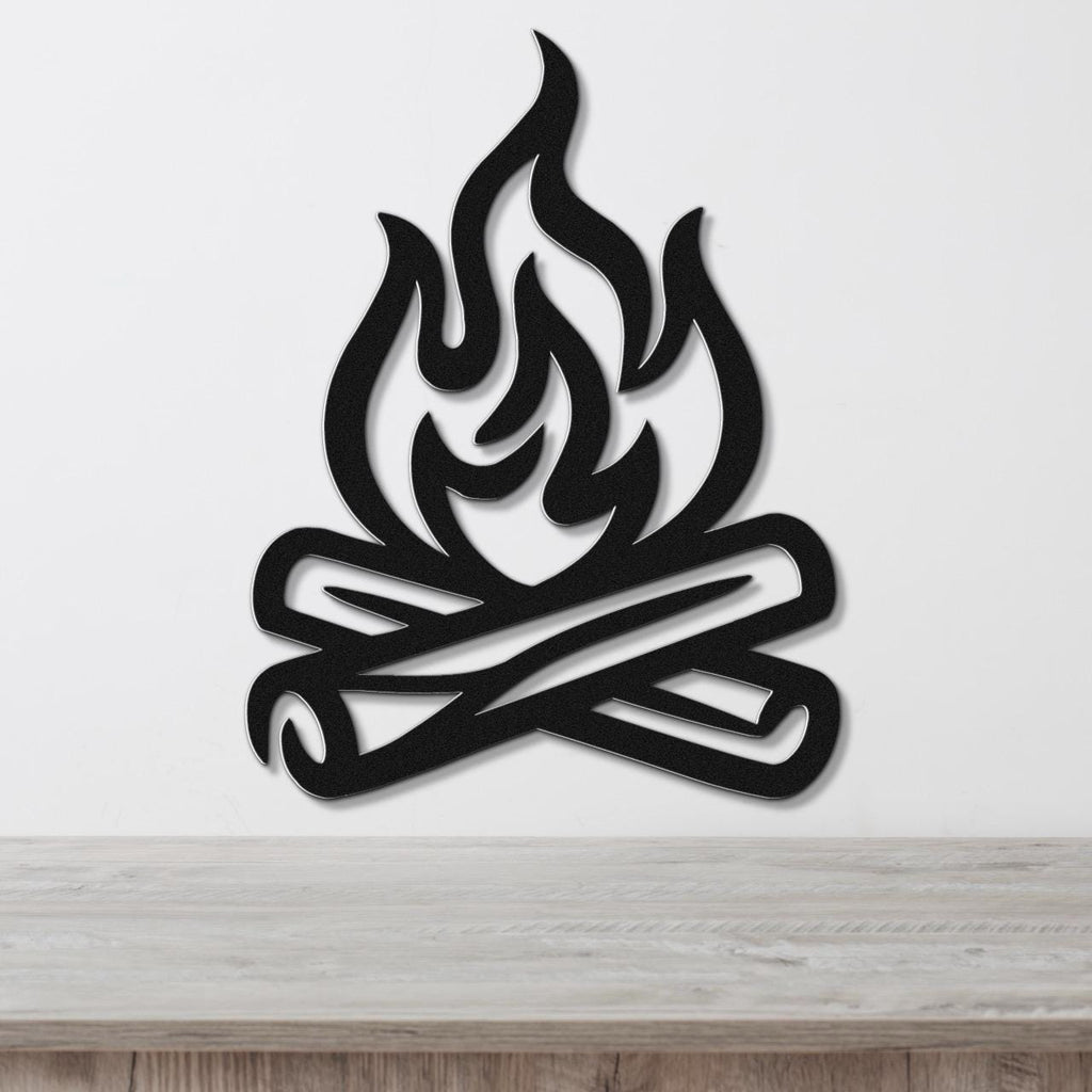 Black metal wall art in a campfire silhouette design with crossed logs and flame cutouts, mounted on a white wall above a wooden surface.