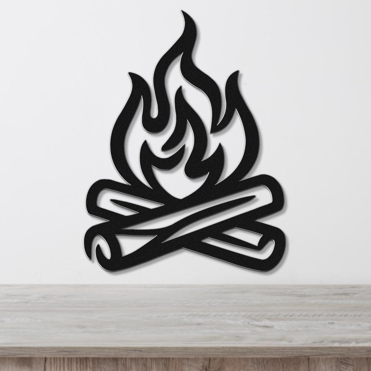 Black metal wall art in a campfire silhouette design with crossed logs and flame cutouts, mounted on a white wall above a wooden surface.