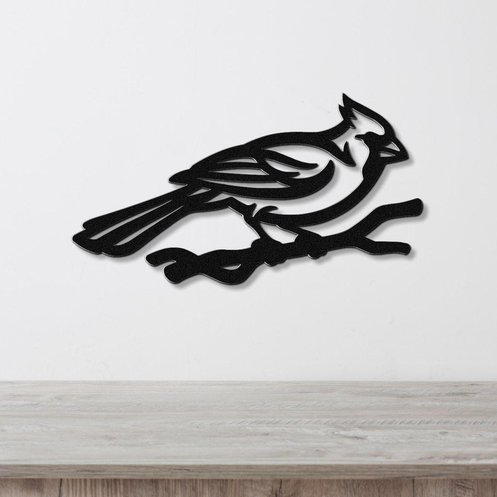 Black metal cardinal wall art in silhouette, perched on a branch with internal cutout details, mounted on a white wall above a light wood surface.