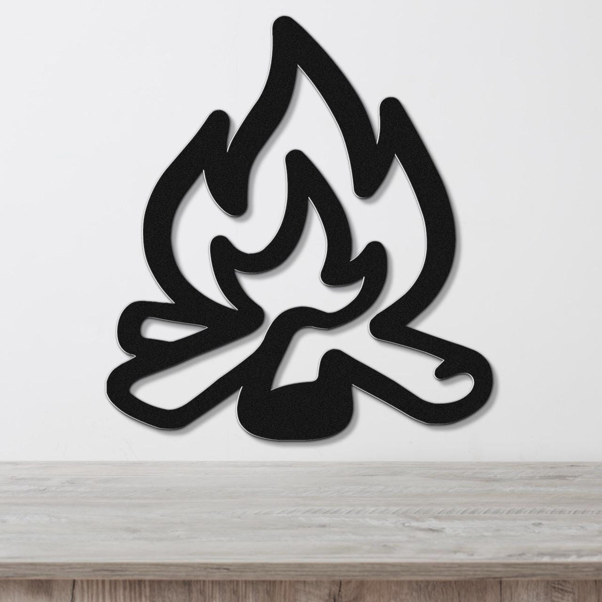 Black metal wall art in a flame silhouette design with cutout logs, mounted on a white wall above a wooden surface.