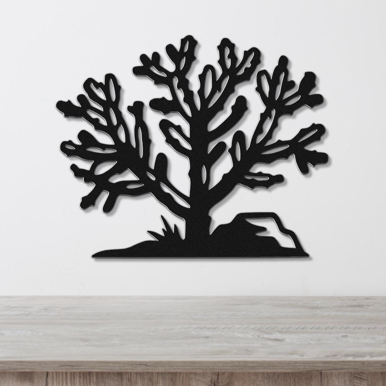 Black metal wall art cutout of a cholla cactus with branching arms and a rock base, mounted on a white wall above a light wood surface.